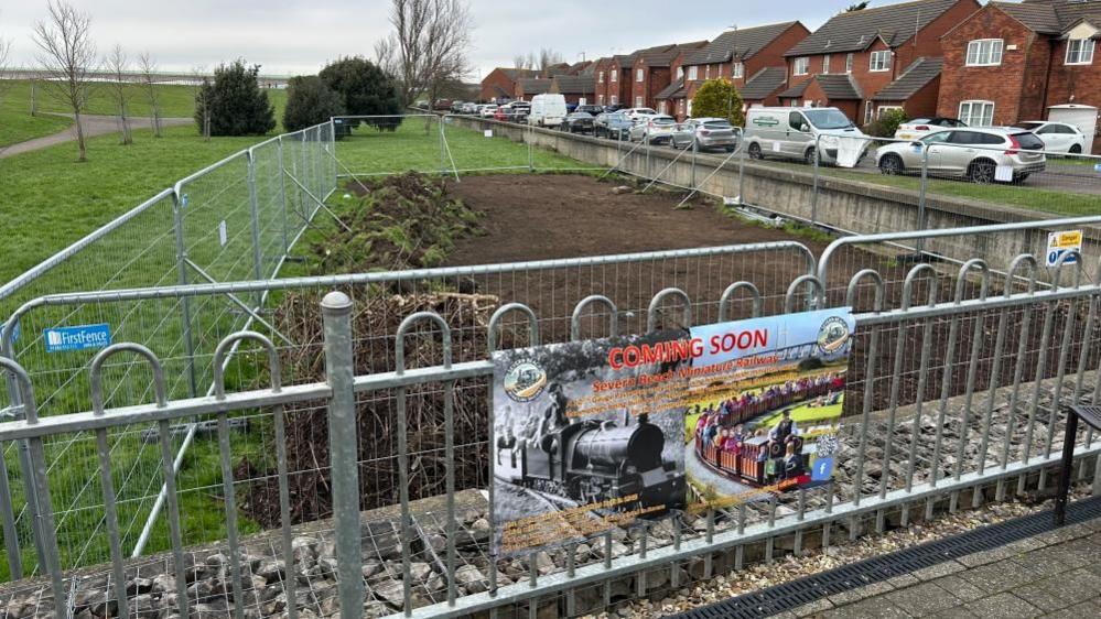 A fenced off field area, with grass partially removed, and a banner saying "coming soon, Severn Beach Miniature Railway" hung on the silver fencing.