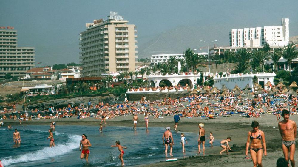 Hundreds of people in swimwear are on a beach in Tenerife, with palm trees and blocks of flats in the background.