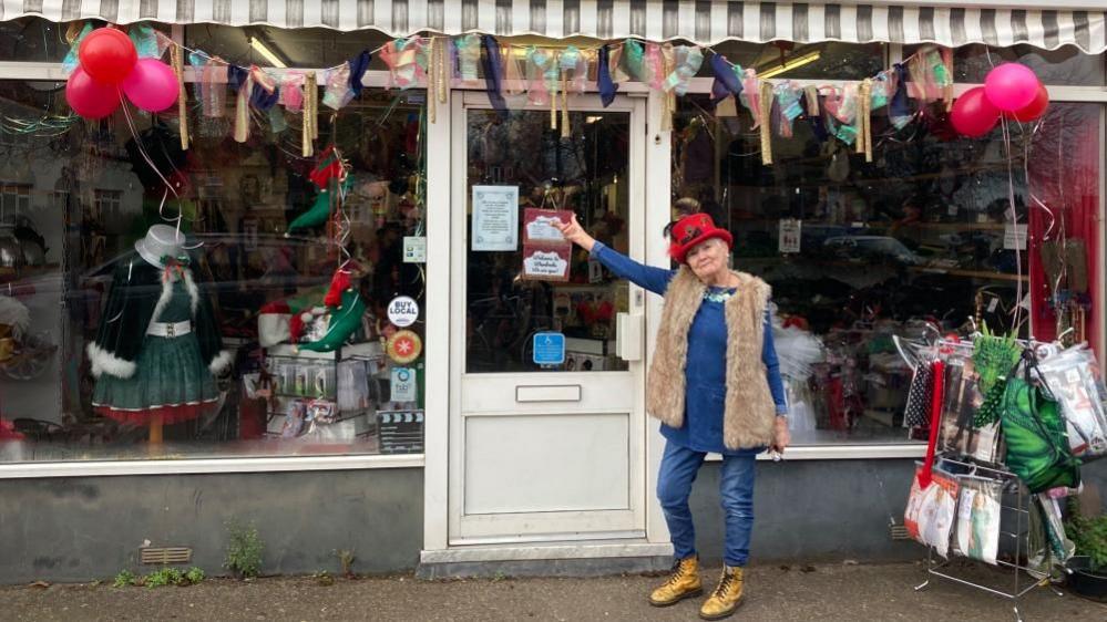 A woman wearing a red hat standing in front of the Wardrobe costume shop. Various costumes can be seen in through the shop window. Balloons and bunting have been attached to the shop front.