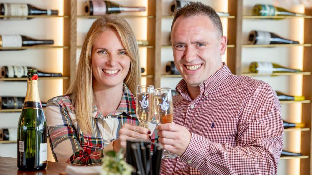 Luke Harris (on the right, a man with short brown hair and smiling looking into the camera, wearing a red and white chequered shirt) and Alison Coke (on the left, a woman with blonde hair looking directly into the camera and smiling, wearing a patterned dress consisting of white, green and red) each holding a glass of champagne and celebrating their win