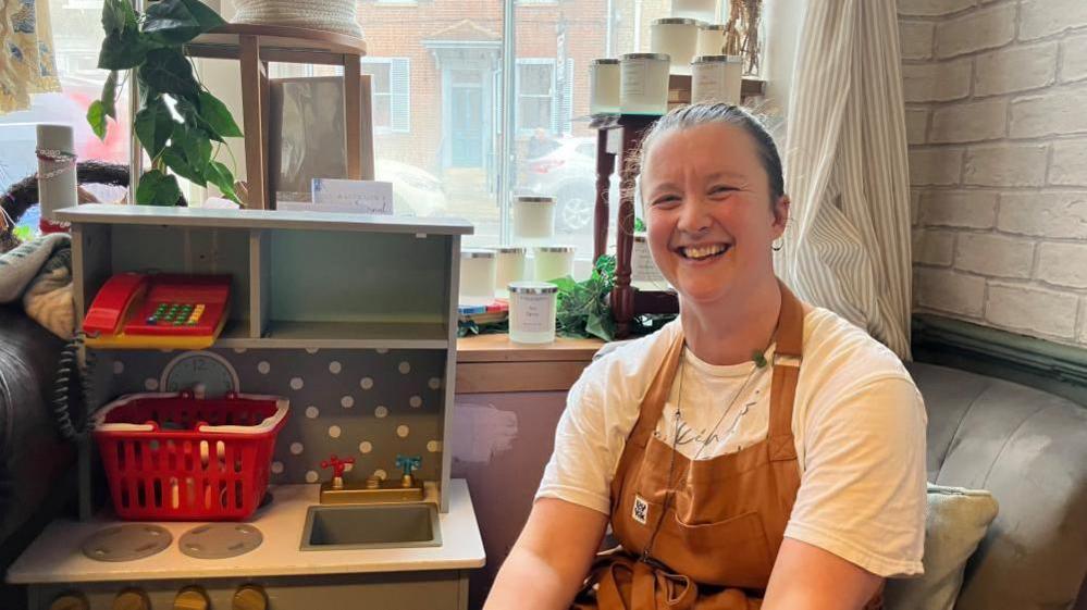 A woman is  seated indoors beside a small toy kitchen set with a red basket and play sink. She is wearing a white T-shirt layered with a brown apron, over it.  She is smiling at the camera and has her hair tied back. Behind her are candles on a stand, a window with light curtains, and brick walls.