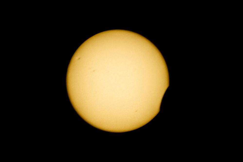 A yellow circle – the Sun – sits against a black sky. There is a small crescent “bite” of black removed from the lower right corner as the Moon moves across it

