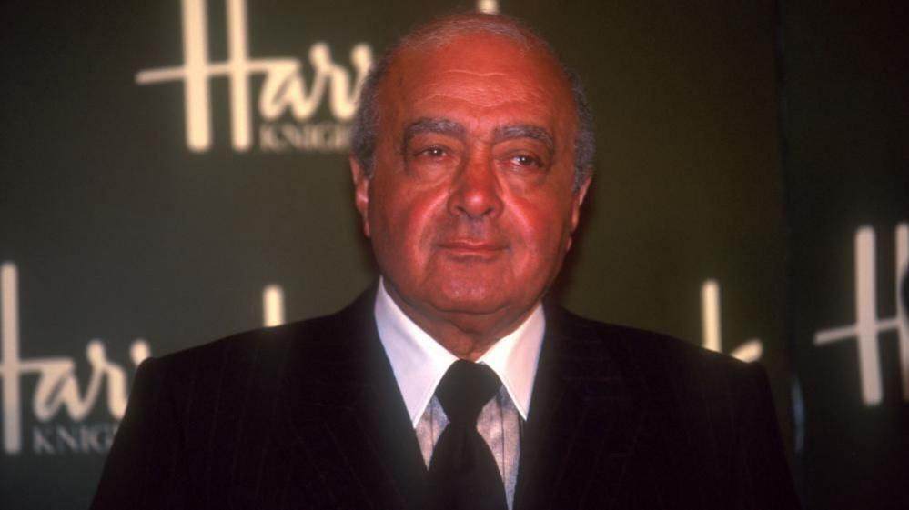 Fayed in a dark suit in front of a Harrods sign