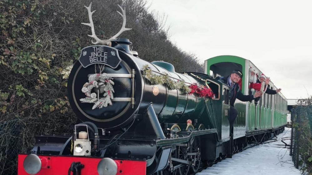 A train conductor, a man in a Santa costume and another man stand leaning out of a miniature train, waving. There is snow on the ground. 