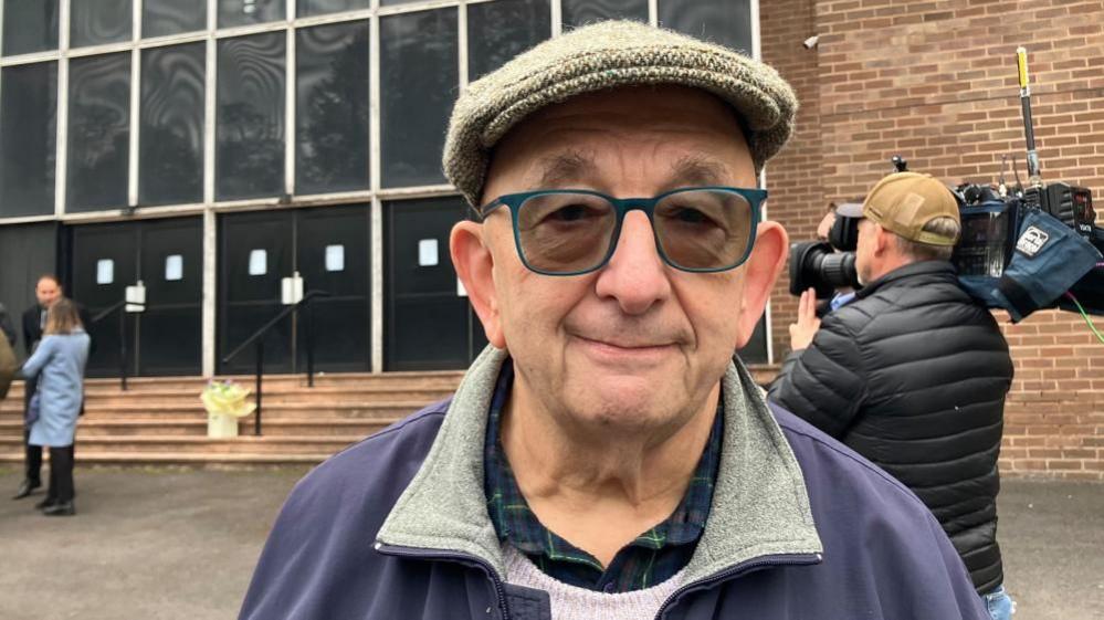 An older man wears a flat wool hat, tinted glasses and stands in front of a large brick building.