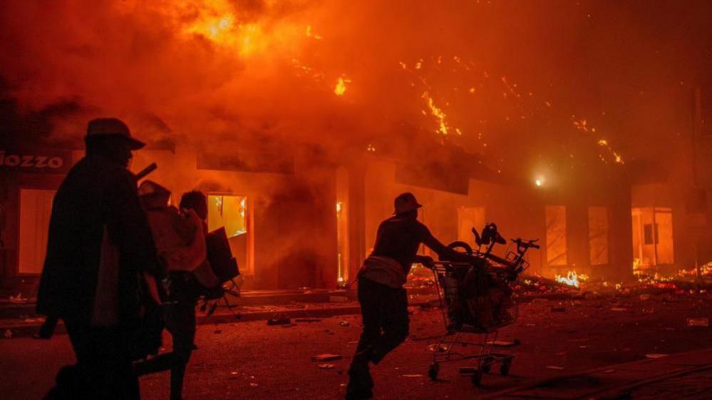 People with their arms full of things, and pushing a shopping trolley loaded with items, run past buildings that are on fire with huge flames licking out of them.