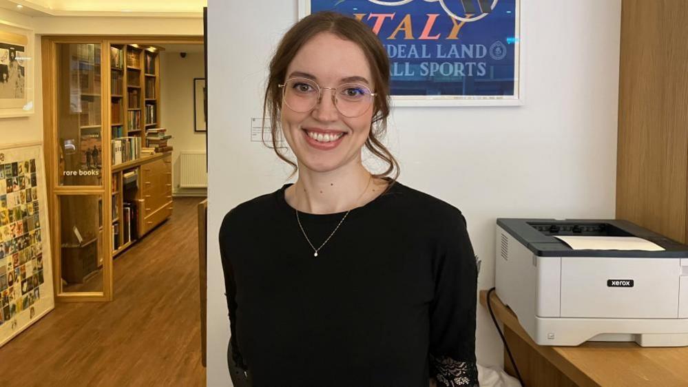 A young woman stands in a bookshop, with a printer beside her on the desk. She has brown hair worn up, with large, silver-rimmed glasses and a silver necklace, and wears a black top.