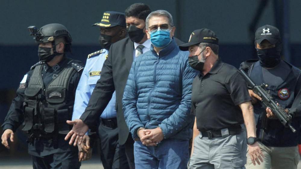 Honduras former President Juan Orlando Hernandez is pictured being escorted by authorities as he walks towards a plane for his extradition to the United States in 2022. Around him are five officials, two of whom are armed. He is handcuffed and wearing a face mask, sunglasses and a blue puffer jacket and a pair of blue jeans. 