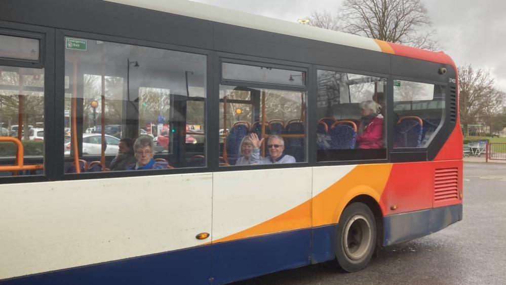 A white, orange and red Stagecoach bus. Nick and Kerstin are sitting in the last seats before the tiered section at the rear of the bus. They are waving to the camera. There is an elderly lady in a seat in front of them, and another in a seat behind them.