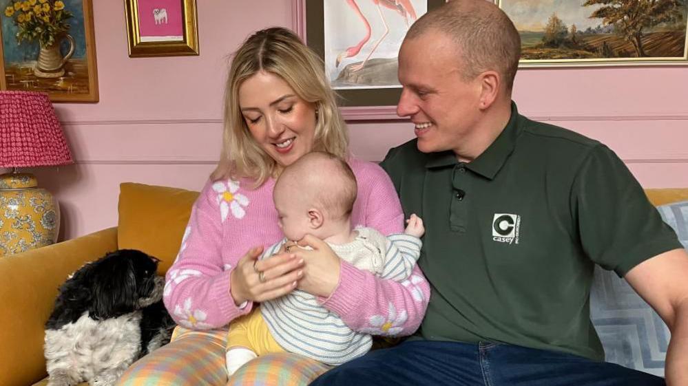 Jessica and Rick Sowerby sit on a sofa with their baby son on Mrs Sowerby's knee. Mrs Sowerby has bobbed blonde hair and is wearing a pink jumper with a daisy pattern. Mr Sowerby wears a green polo shirt. Both are smiling and looking at their son, who wears a grey and white striped jumper, yellow trousers and white socks. The couple's dog Merla can be seen on the sofa beside them.