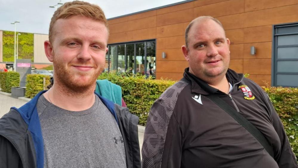 Two men look at the camera, smiling. One has short ginger hair and is wearing a grey t-shirt with a black jacket unzipped down the middle. He is standing next to a man with a balding head and wearing a black sport's top with a bag across his chest.