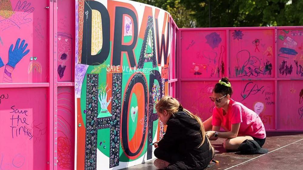 Two artists sit on the ground and paint on a large piece of artwork that is hanging on a bright pink wall.