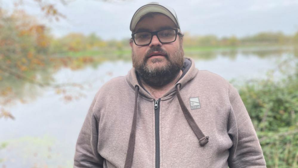 Craig Parkins, standing outside, wearing a cap, with glasses on, wearing a pale hoodie, with a beard. He is standing in front of a lake, with grass to the side. 