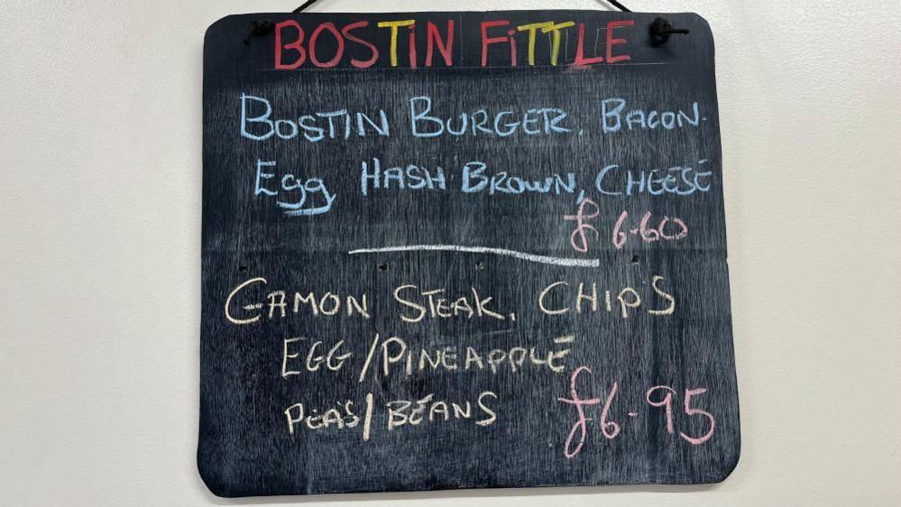 A picture of a small blackboard hanging on a white wall. In red and yellow letters at the top it reads "Bostin Fittle". Underneath, in blue letters, it says "Bostin burger, bacon, egg, hash brown, cheese", with the price written in pink letters - £6.60. Below, in yellow letters, it says "gammon steak, chips, egg/pineapple, peas/beans", and the price again in pink letters - £6.95.