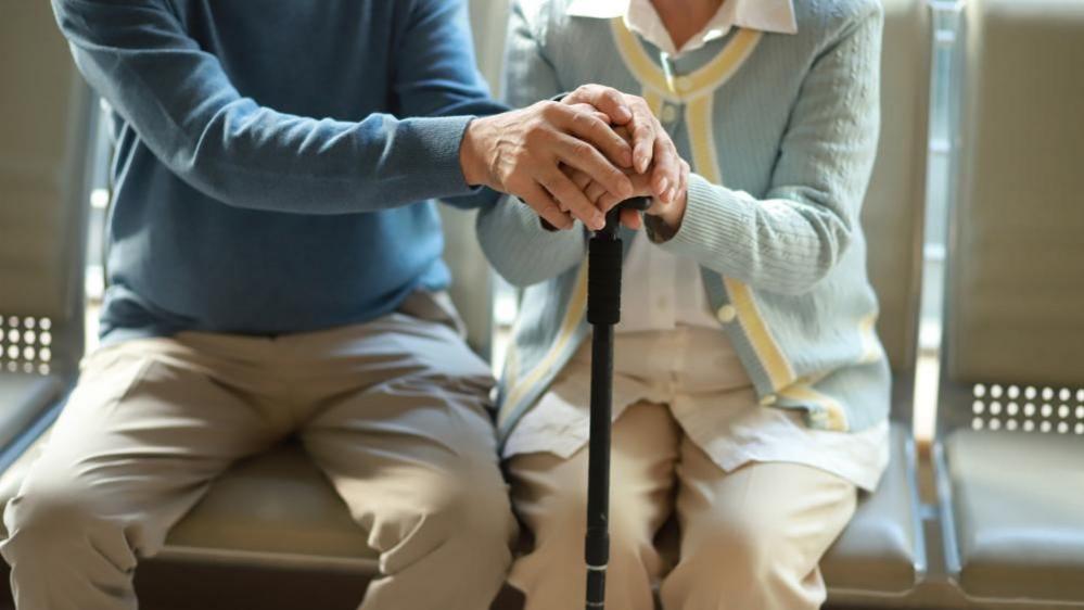 Elderly couple on a row of seats. Their faces are not shown but a man and a woman, both wearing cream-coloured trousers and blue sweaters, can be seen sitting next to each other on a row of seats. The woman is holding a walking stick 