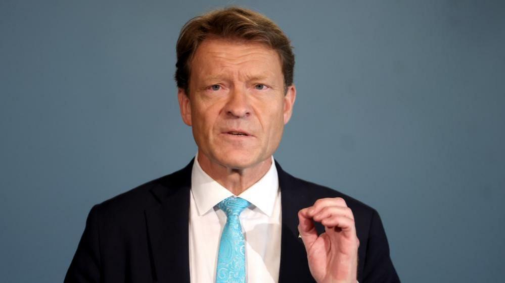 Richard Tice speaks to a crowd, wearing a black suit and white shirt with a blue tie