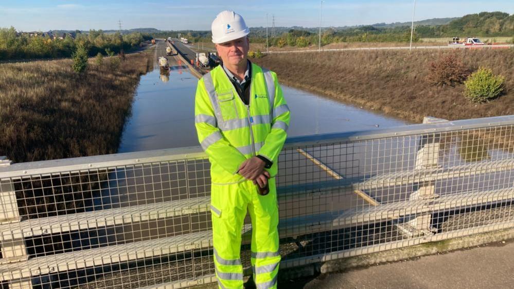 Regional director of National Highways, Martin Fellows, standing on a bridge, looking at the camera, with his arms crossed. He is wearing a hard hat and high viz clothing. The road behind him is flooded.