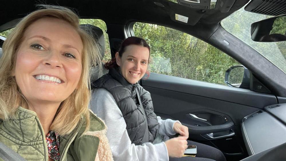 Two women sitting in the front of a car. They are both smiling and looking into the camera. One is blonde one and is brunette