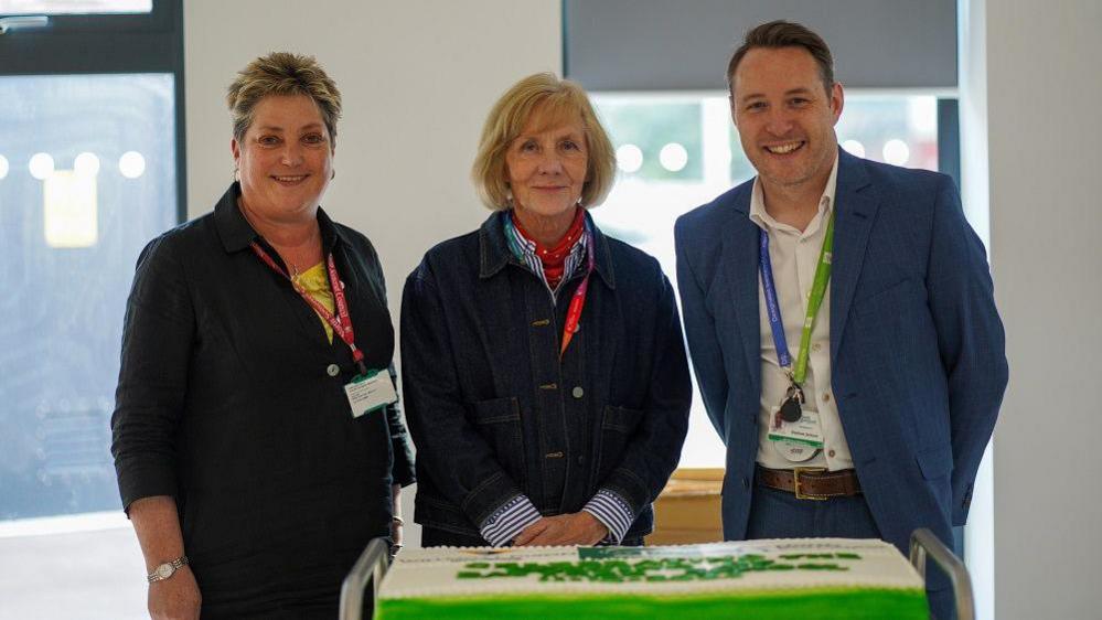 Three people smiling at the camera standing behind the cake on a trolley