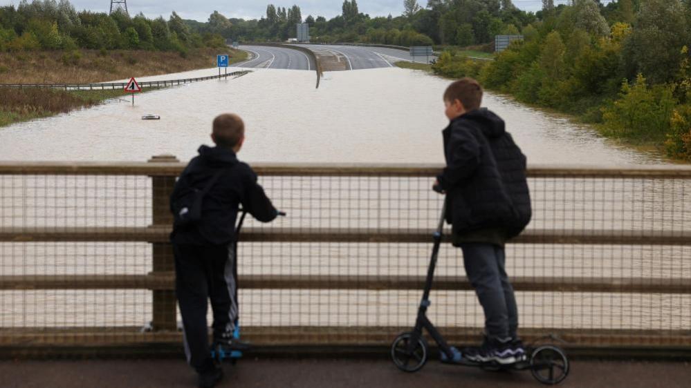 Two boys on scooters stood on a road bridge looking at the flood water