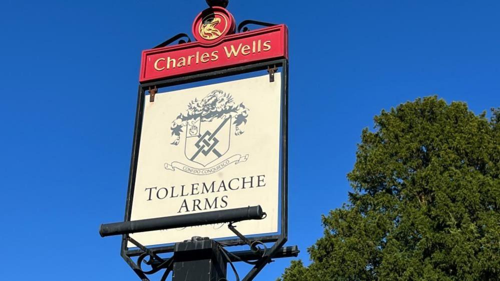 Pub sign on top of a post for the Tollemache Arms showing a coat of arms on a white background, and red Charles Wells section above the swinging bit of the sign.