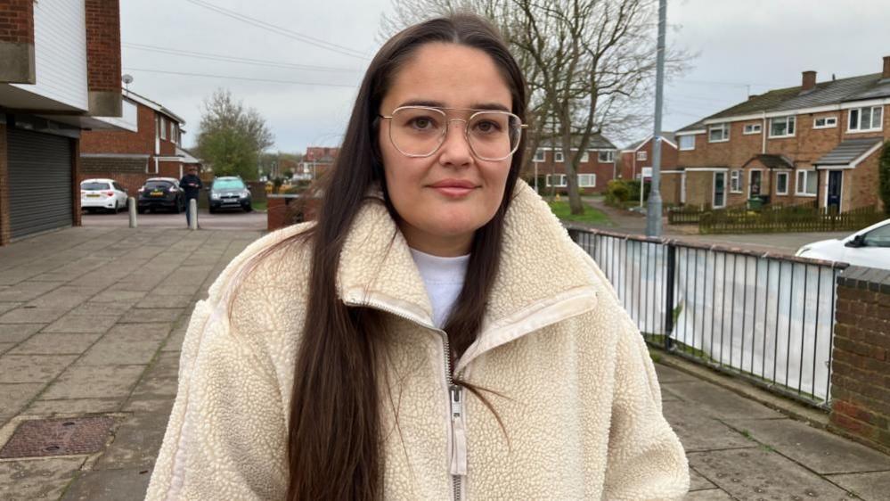 Chelsea Moncur, standing in a street with cars and houses behind her. She has long, straight hair, with some of it hanging down the front of her coat. She is wearing glasses and a white top and cream fluffy coat. She is looking straight at the camera.