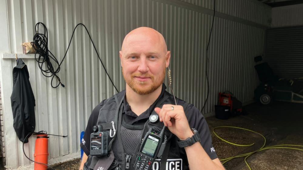 A police officer is standing in uniform in front of a bay in a car wash. He is wearing a stab vest with a bodyworn camera on one side and a radio on the other. He is wearing an earpiece, connected to his radio. 