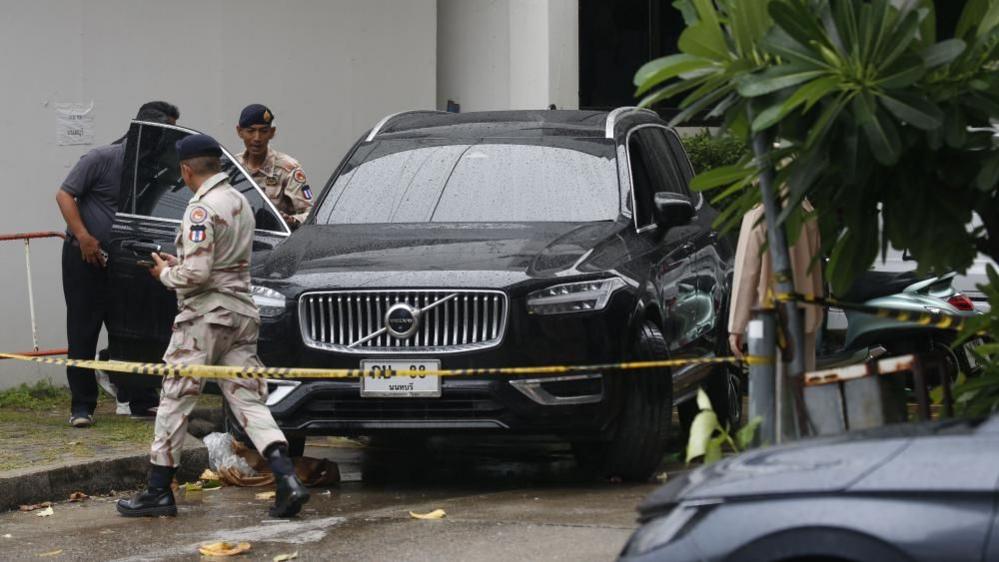 Police officers inspect a black car surrounded by police tape