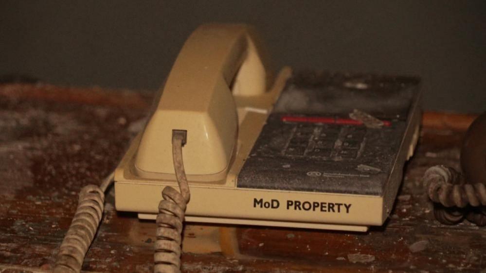 A cream-coloured telephone, with dark grey keyboard pane, with the receiver and wire visibly dirty. It is marked MoD Property and is on a desk with dirt and detritus caused by the ceiling becoming damaged.