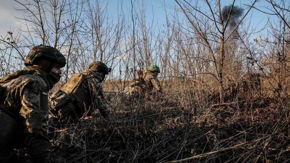 Three Ukrainian soldiers crawling through bushes with an explosion in the distance