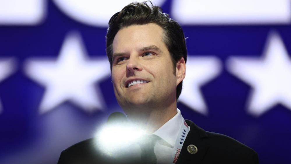 Wearing a suit with his congressional pin, Matt Gaetz stands at a microphone on a stage with a backdrop of white stars on a blue background. 