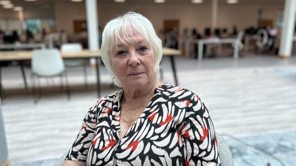 Danielle Stone with short blond hair, wearing a white, black and red top and sitting in a modern hall with tables and plastic chairs.