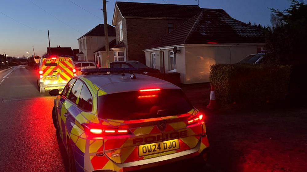 A police car and a police van are parked outside houses at the side of a road. It is just before sunrise and the vehicles' lights are on