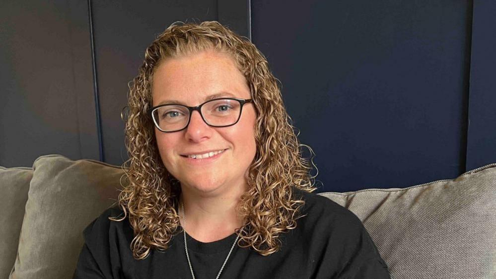 Aimee Tilley looking at the camera and smiling. She is wearing a black T-shirt and sitting on a grey sofa