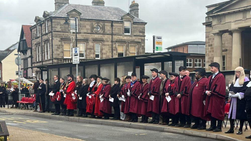 A row of people in burgundy coats and black hats along the side of a road with large brick buildings behind them