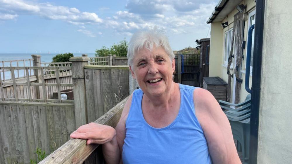 Carol Boyes has medium length white hair. She is wearing a blue vest top, and has her right hand on the garden fence. Behind her is more fencing, her house to the right of the image, and the sea visible to the left.