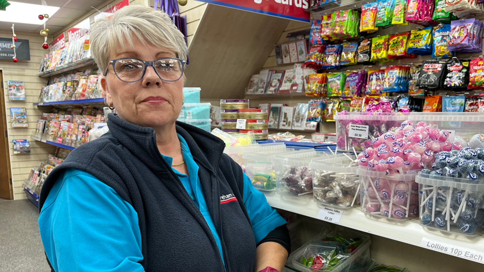 Carol Clarke stands beside a shop counter with piles of sweets in clear boxes stacked on its surface. She looks serious and directly at the camera. She has short grey hair and blue-rimmed glasses. She is wearing a blue polo shirt and dark blue gilet. Behind her is a wall stack displaying magazines.