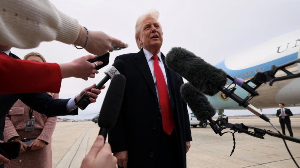 Donald Trump in long dark coat with red tie, surrounded by microphones and hands in front of Air Force One 