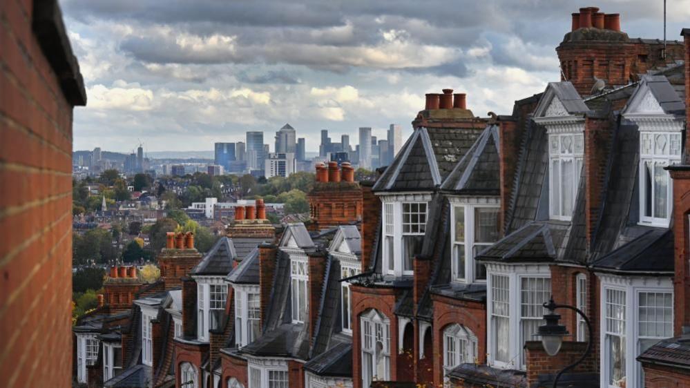 A row of houses along a street on a hill in London, with the skyscrapers and tower blocks of Canary Wharf in the background.
