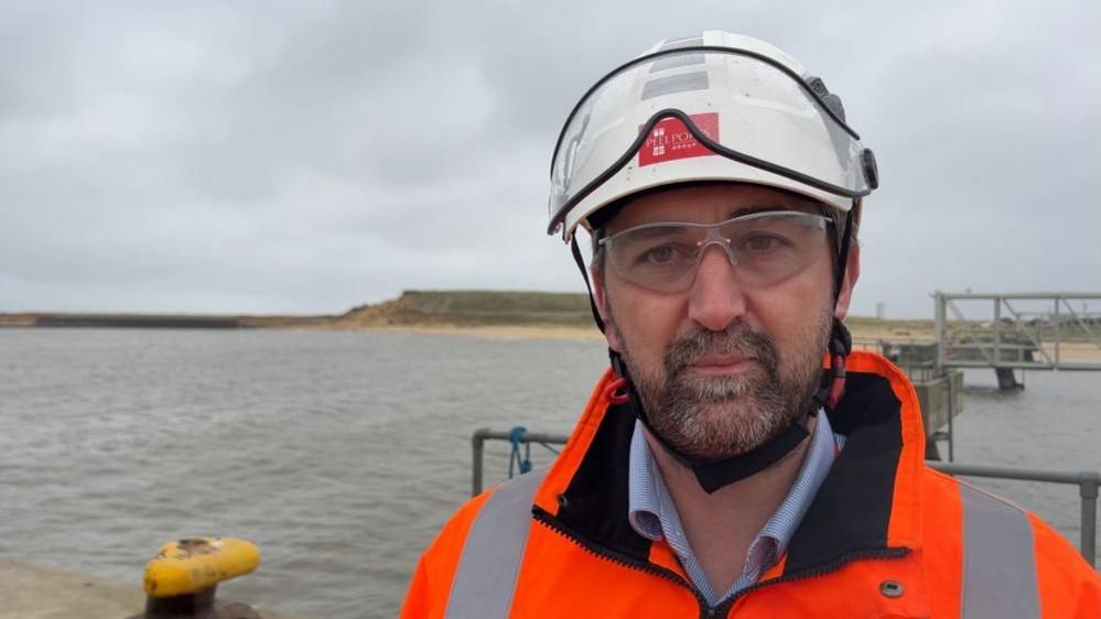 Richard Goffin is wearing a white hard hat, with visor, and safety glasses, and a orange high visibility coat. He is standing near the outer harbour quayside west terminal, with the undeveloped southern terminal behind him. There are port structures over his left shoulder.