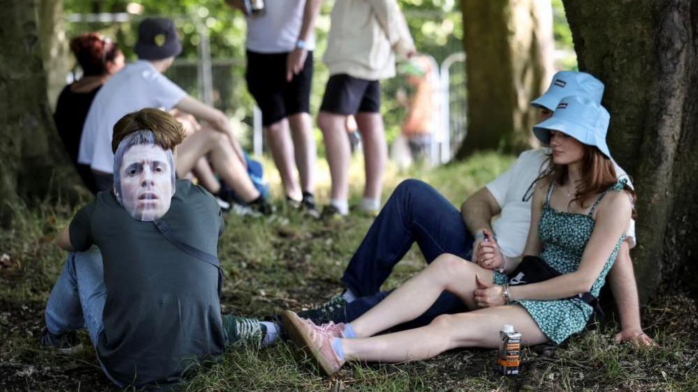 Three people sit in the shade beneath trees. Two wear bucket hats and one has a Liam Gallagher facemask behind his head. Another group can be seen sitting in the shade behind them.