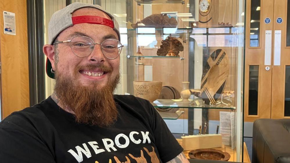 Connor Parker is wearing a baseball cap backwards with the strap across his head. It's a sand coloured cap with read strap. He is wearing a black t-shirt with "We rock" on the front. He is smiling at the camera, wearing glasses and has a nose ring.