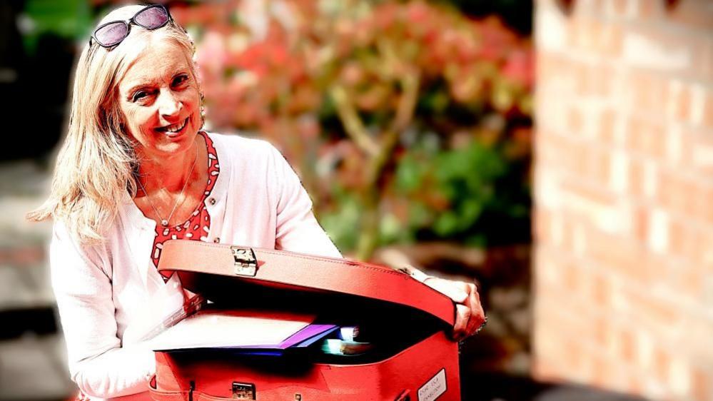 Woman wearing white cardigan and red dress smiling next to red case overflowing with papers and files