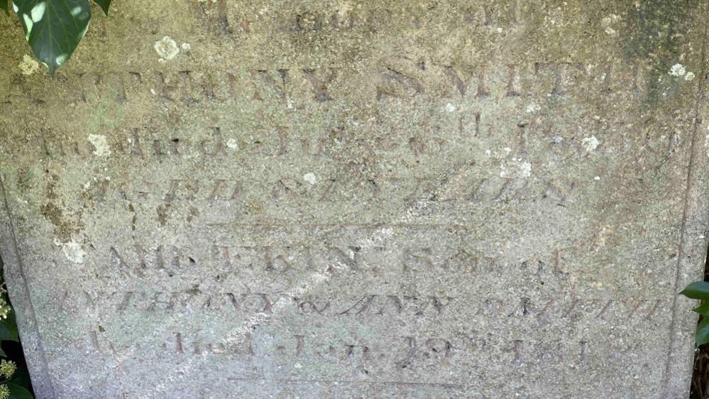 The gravestone of Anthony Smith with very faint words etched on it. There are leaves around it. 