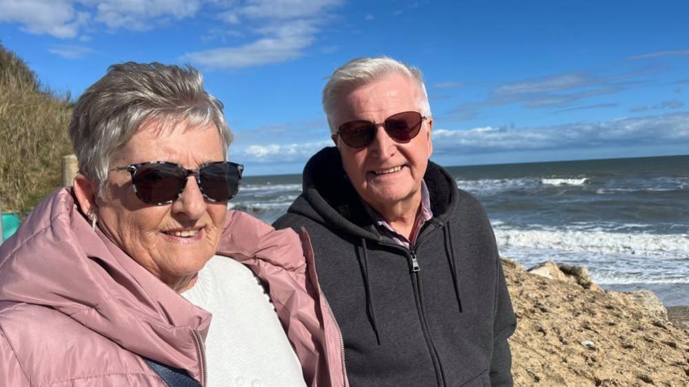 Diana Holmes, left, wearing a pink jacket and her husband Malcolm, right, in a black hooded top, sitting near the beach in Hemsby