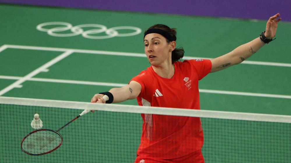 Kirsty Gilmour is playing badminton with her racket outstretched to hit a shuttlecock near the next. She wears a red uniform with the Team GB logo, as well as a black headband and black sweatbands on her wrists.