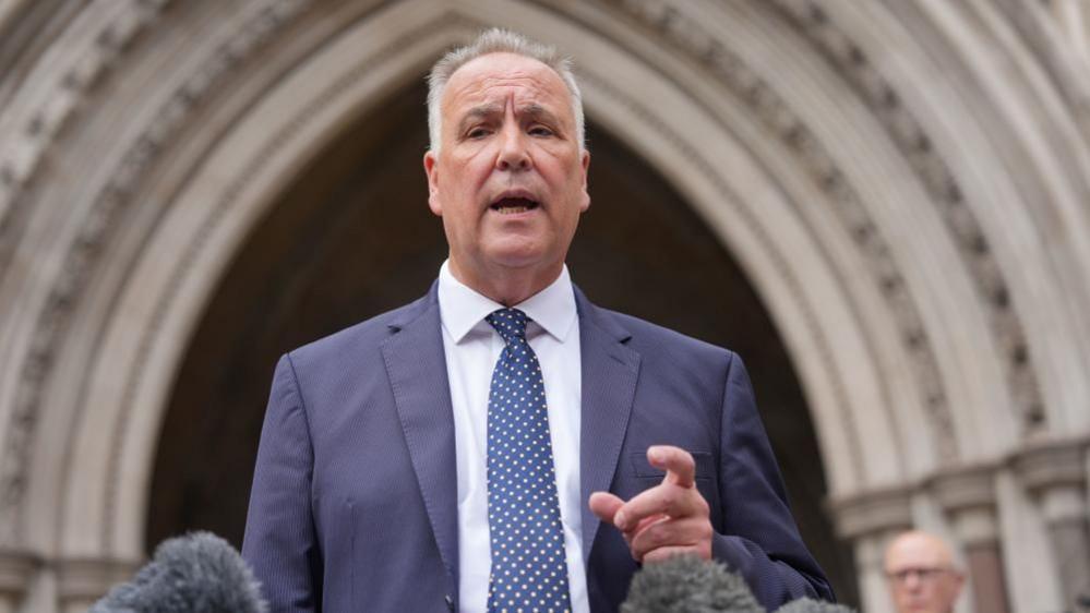 Chris Whitbread, wearing a blue suit, white shirt, and blue and white spotted tie, outside the the Royal Courts of Justice, talking into microphones, with his leg hand raised. He has short grey hair. Another man is behind him, and an archway is behind him. 