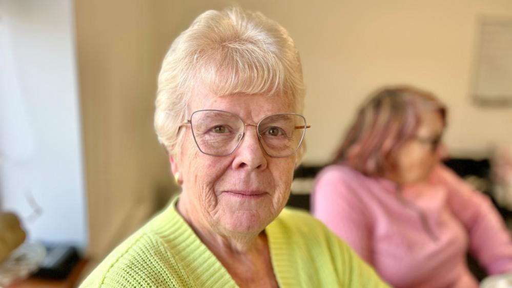 Sandra Taylor is sitting at a table in the community center wearing a bright lime-green knitted sweater and glasses. Sandra is smiling at the camera. In the background, another person is blurred but you can see she is wearing a pink top and has shoulder-length hair with reddish tones. 