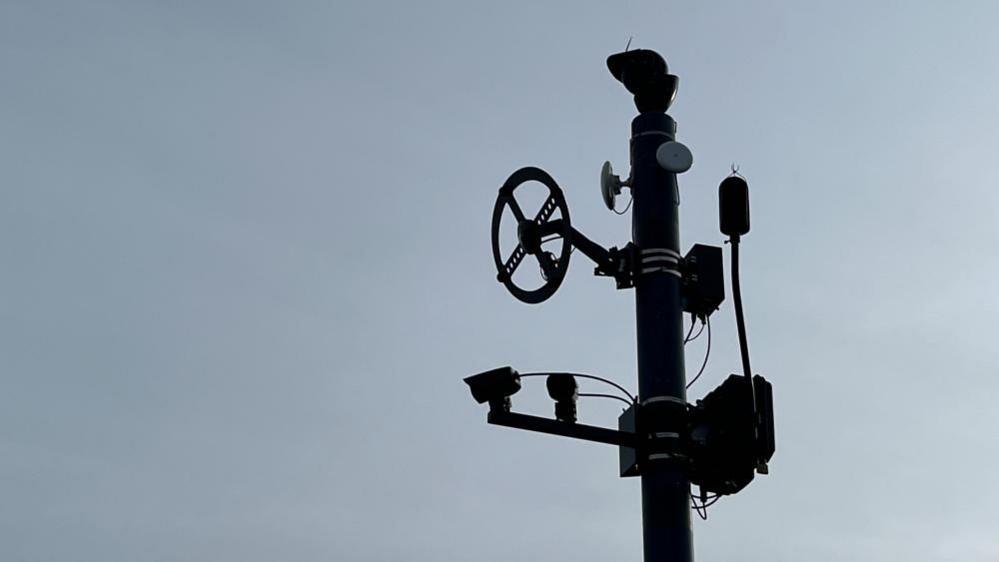 A sound camera which uses cameras and sound recording devices to capture evidence on antisocial driving. It shows various aerials and cameras on a pole, in silhouette.