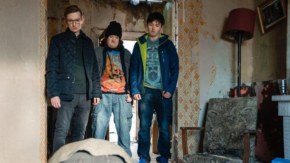 Callum (Ryan Dylan), Jordan (Rian Lennon) and Lorcan (Lee Dobbin)  pictured in an old-fashioned, rundown rural living room with torn 1970s style wallpaper and damaged furtniture. All three men are pictured in scruffy clothing, dirty baggy jeans and hoodies